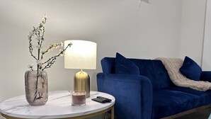 Una sala de estar moderna con una mesa redonda blanca, un sofá azul, una lámpara dorada, un florero con ramas y una vela.