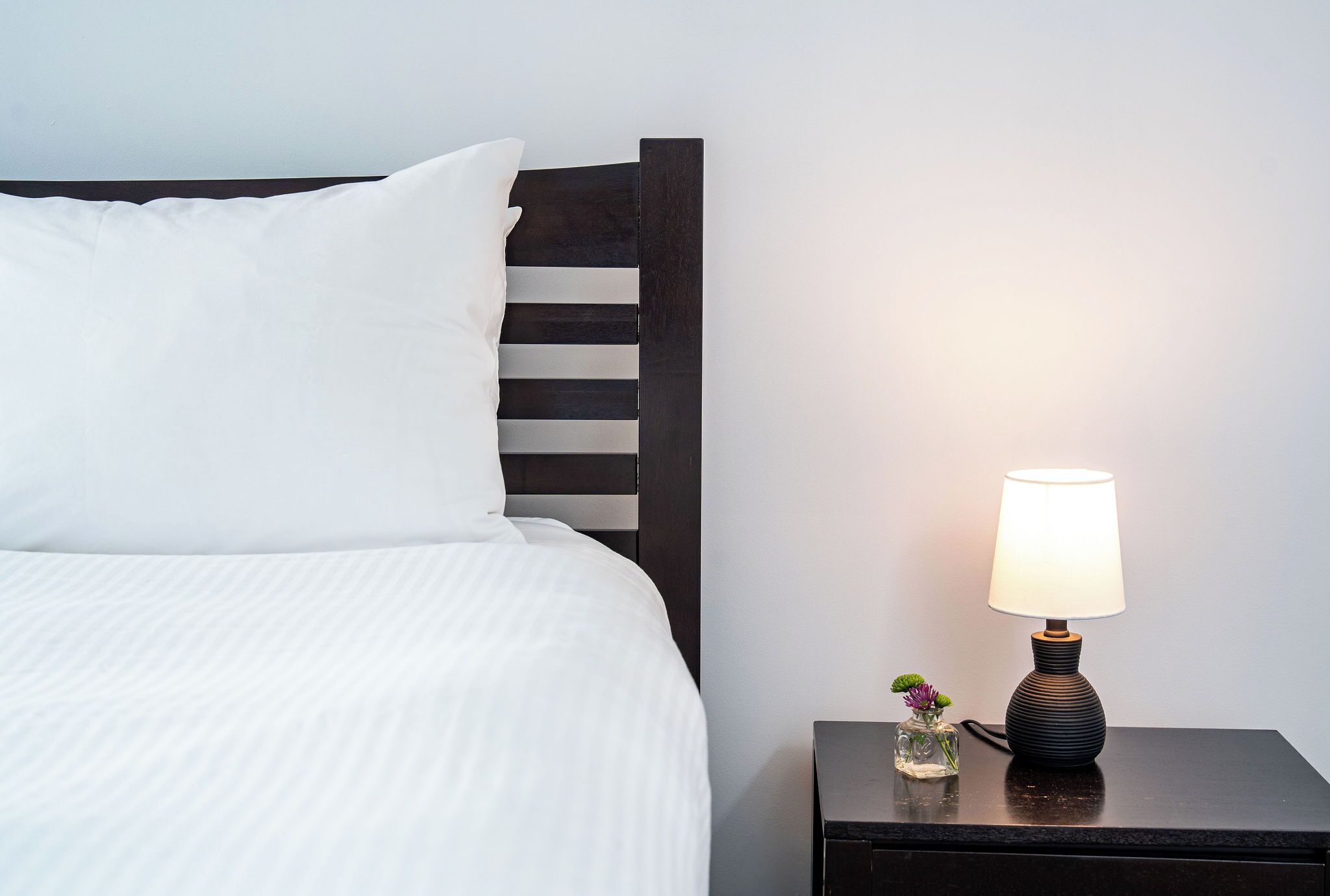 Een netjes opgemaakt bed met witte beddengoed, een donker houten hoofdbord en een nachtkastje met een lamp en een kleine vaas met bloemen.