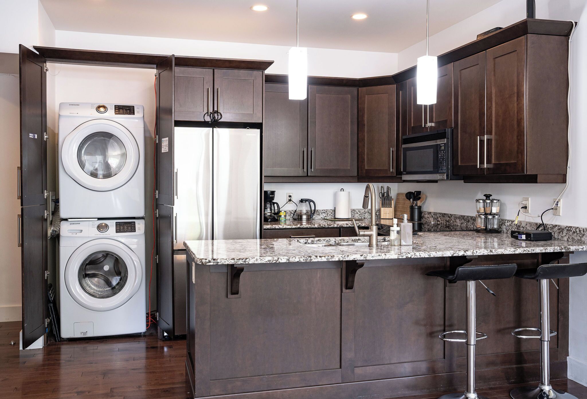 A kitchen with dark wood cabinets, a built-in microwave, a stainless steel refrigerator, a double washer, and a granite countertop.