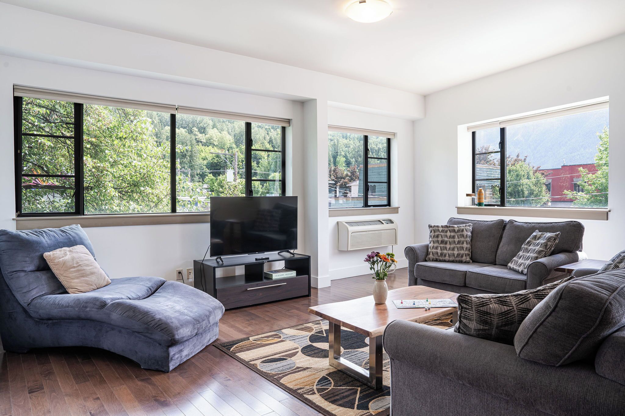 A modern living room with a flat-screen TV, two grey sofas, a coffee table, and large windows with a view of greenery and mountains.