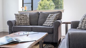 Un sofá gris con cojines estampados, una mesa de centro de madera con juegos de mesa y una ventana con vista a la montaña.