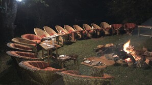 An outdoor dining area with wicker chairs and tables, a fire pit, and a string of lights. - Explore Mara Camp (Maasai Mara)