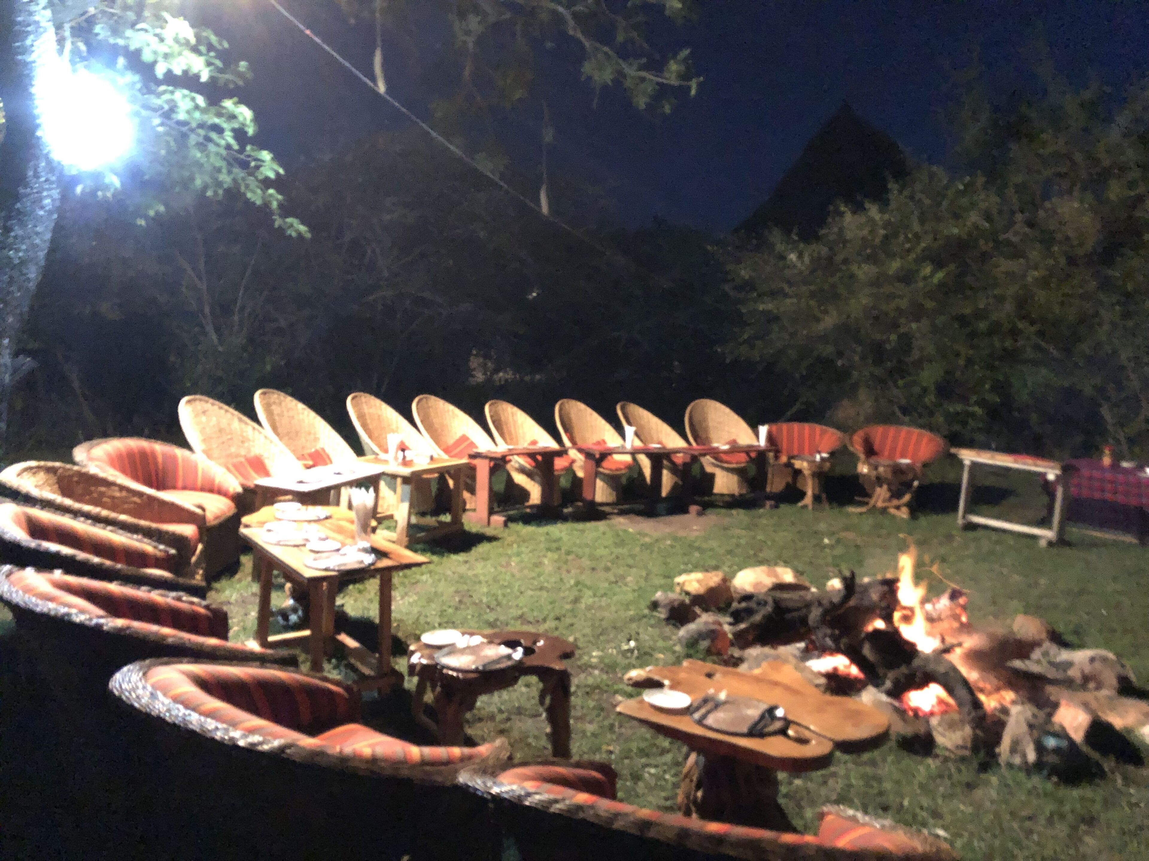 Un espace extérieur aménagé avec des chaises et des tables en osier, un foyer et une table recouverte d’une nappe à carreaux.
