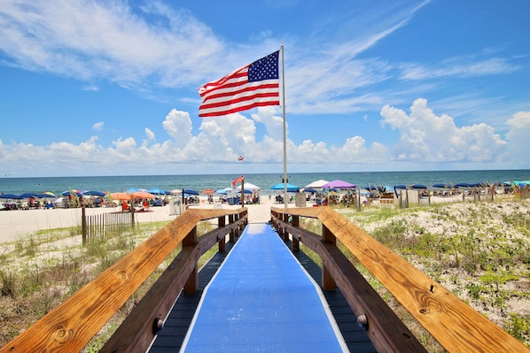A wooden boardwalk leading to a beach with umbrellas and a flag.