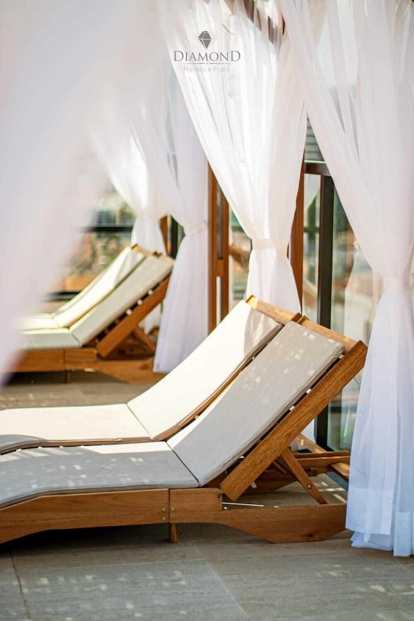 Una fila de sillones de madera con cojines blancos, dispuestos en un espacio al aire libre iluminado por el sol y con cortinas blancas translúcidas.