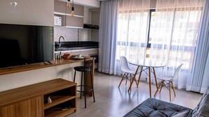 Una sala moderna con un televisor de pantalla plana, un mueble de entretenimiento de madera y un sofá gris. Hay una zona de cocina con encimera negra, armarios blancos y un microondas. Una mesa de comedor con sillas blancas está ubicada cerca de un amplio ventanal con cortinas translúcidas.