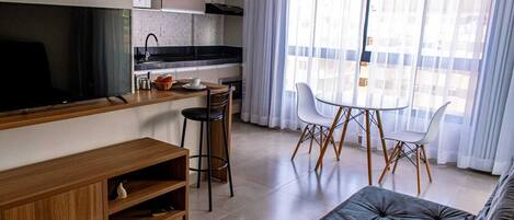 Una sala moderna con un televisor de pantalla plana, un mueble de entretenimiento de madera y un sofá gris. Hay una zona de cocina con encimera negra, armarios blancos y un microondas. Una mesa de comedor con sillas blancas está ubicada cerca de un amplio ventanal con cortinas translúcidas.