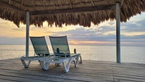 Una terraza de madera con dos sillones de descanso bajo un techo de paja, con vistas al mar al atardecer.
