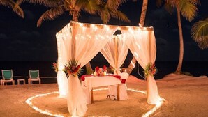 Una carpa frente a la playa con una mesa preparada para dos personas, rodeada de palmeras y luces colgantes.
