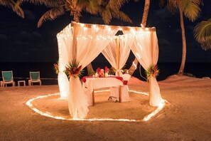 A beachside tent with a table set for two, surrounded by chairs, palm trees, and string lights. - 1BR Casa De Seaclusion @ Sapphire Beach Resort (San Pedro Town)