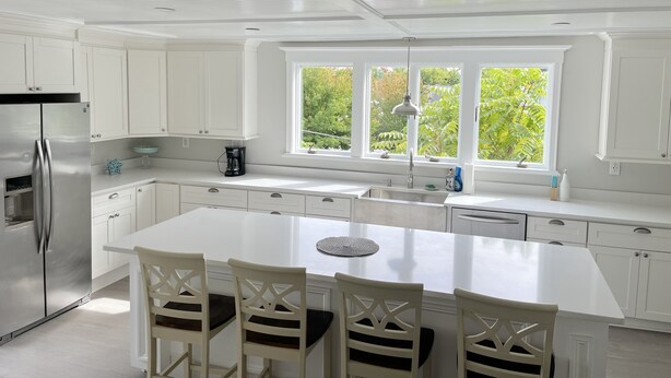 Una cocina moderna con armarios blancos, un refrigerador de acero inoxidable, una isla grande con sillas y un ventanal con vista a la vegetación.
