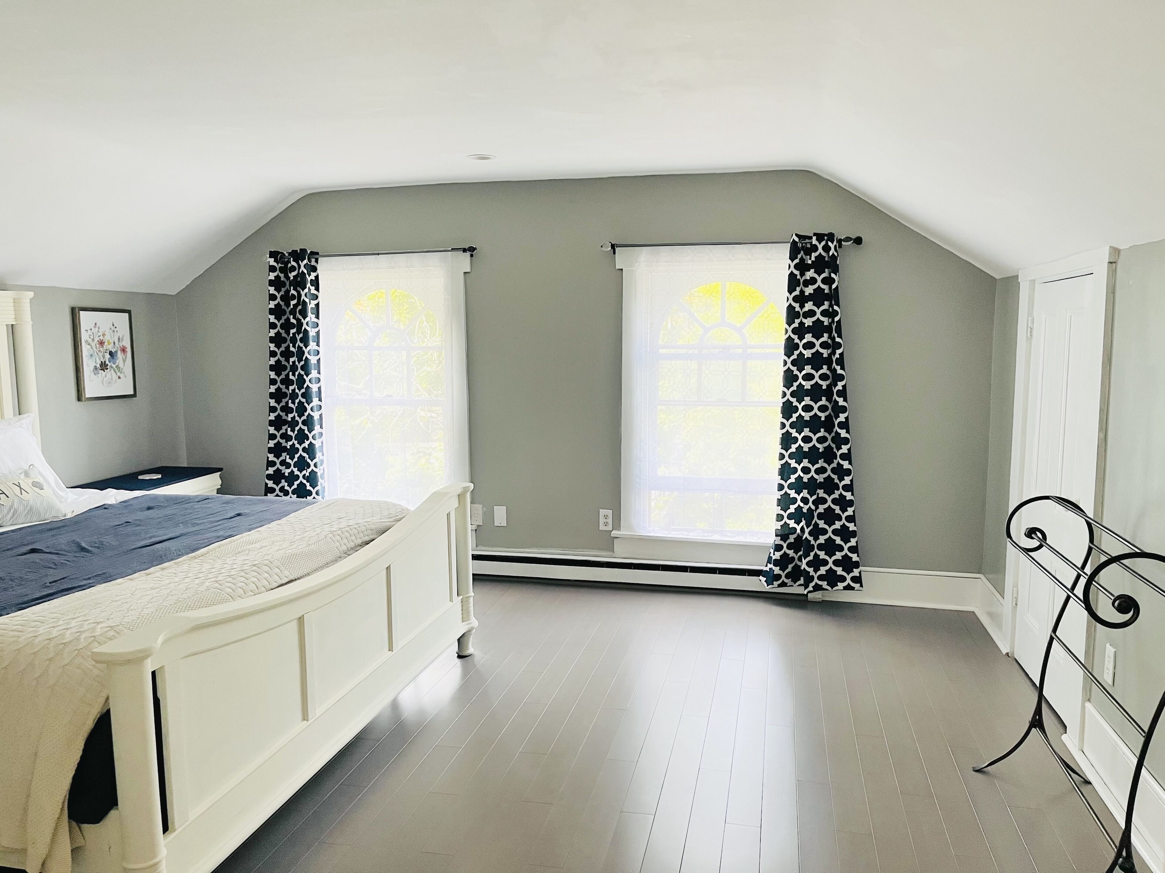 Un dormitorio con cama, dos ventanas con cortinas estampadas, un radiador y un cuadro en la pared.