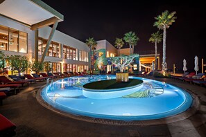 Un área de piscina nocturna con sillones de descanso y palmeras.