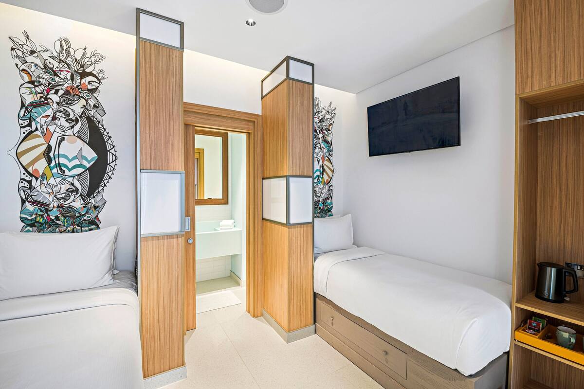 Une chambre d’hôtel avec deux lits simples, un lambris en bois, une grande œuvre d’art abstraite accrochée au mur, un téléviseur à écran plat et une machine à café posée sur la table de chevet.