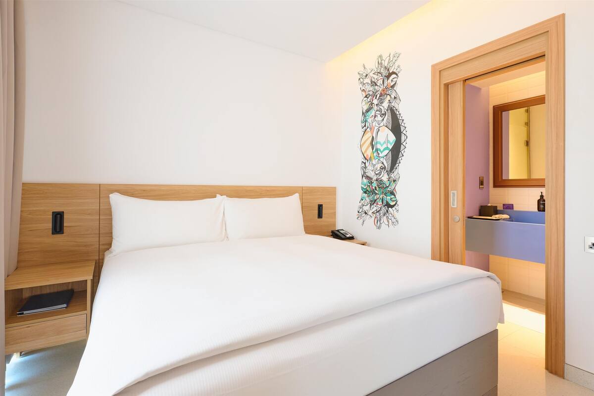 Une chambre d’hôtel moderne avec un grand lit, une tête de lit en bois, une table de chevet avec une lampe, une œuvre d’art accrochée au mur et une salle de bain visible à travers une porte ouverte.