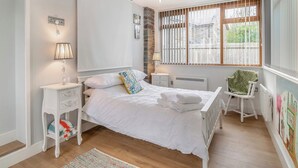 Quarto com cama grande, mesas de cabeceira brancas, radiador, janela com persianas e uma cadeira.