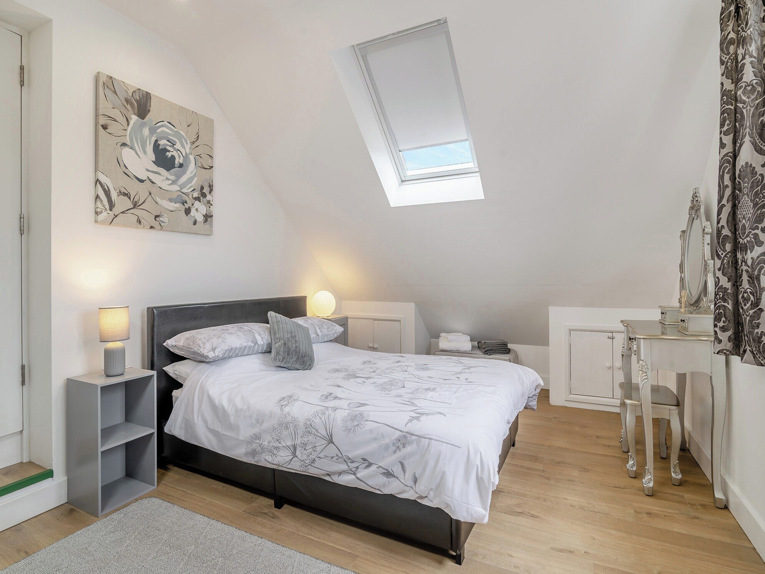 A bedroom with a bed, bedside tables, a desk, a chair, a skylight, and a wall-mounted artwork.