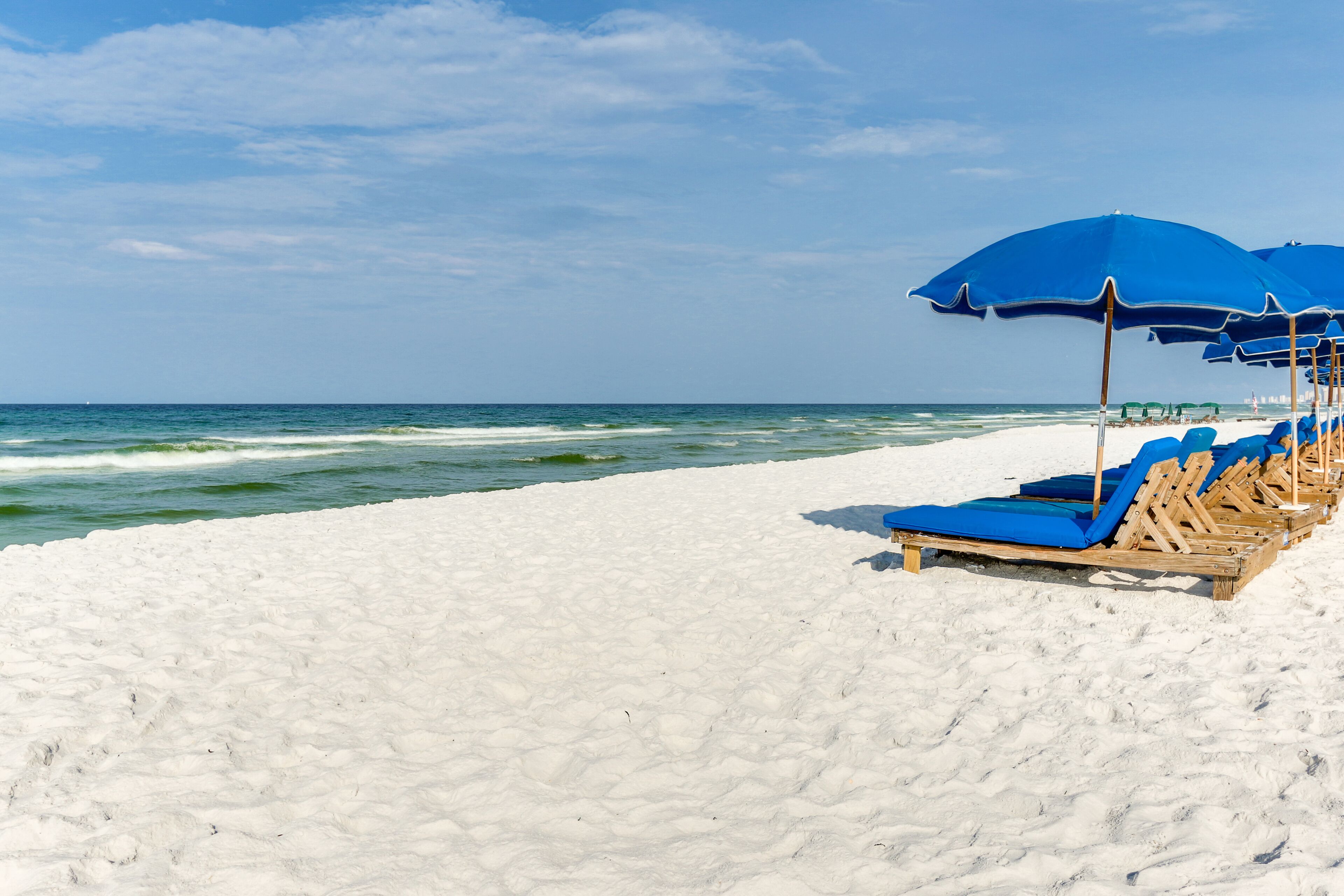 A beach with white sand and ocean waves. There are blue umbrellas and wooden lounge chairs.