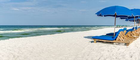 A beach with white sand and ocean waves. There are blue umbrellas and wooden lounge chairs.
