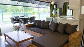 Un espace de vie moderne comprenant un canapé, une table à manger et des chaises, un coin cuisine avec un réfrigérateur, et une grande fenêtre donnant sur un jardin.