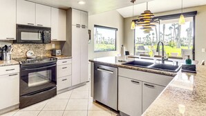 A modern kitchen with a built-in microwave, oven, and dishwasher, a granite countertop, and a large window offering a view of palm trees. - Patio + Resort Amenities: Palm Desert Escape (Palm Desert)