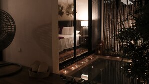 Una habitación tenuemente iluminada con una cama, un ventanal y un pequeño balcón con velas.