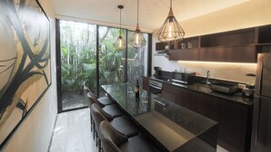 Cocina moderna con una isla grande, taburetes de bar, iluminación colgante y vistas a un jardín tropical.