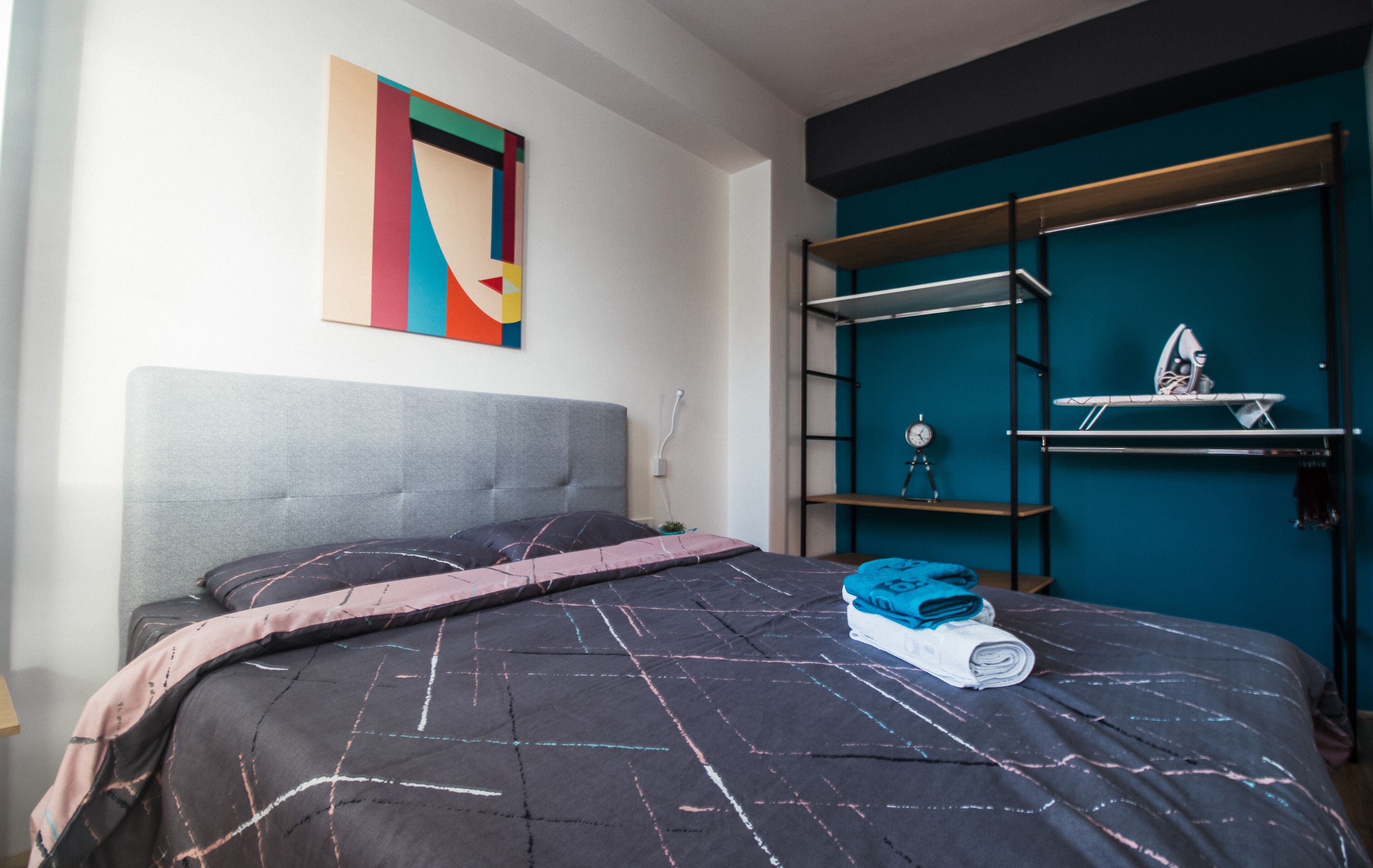 A bedroom with a bed, a colorful abstract painting, a teal accent wall, and a built-in shelving unit with a small ironing board.