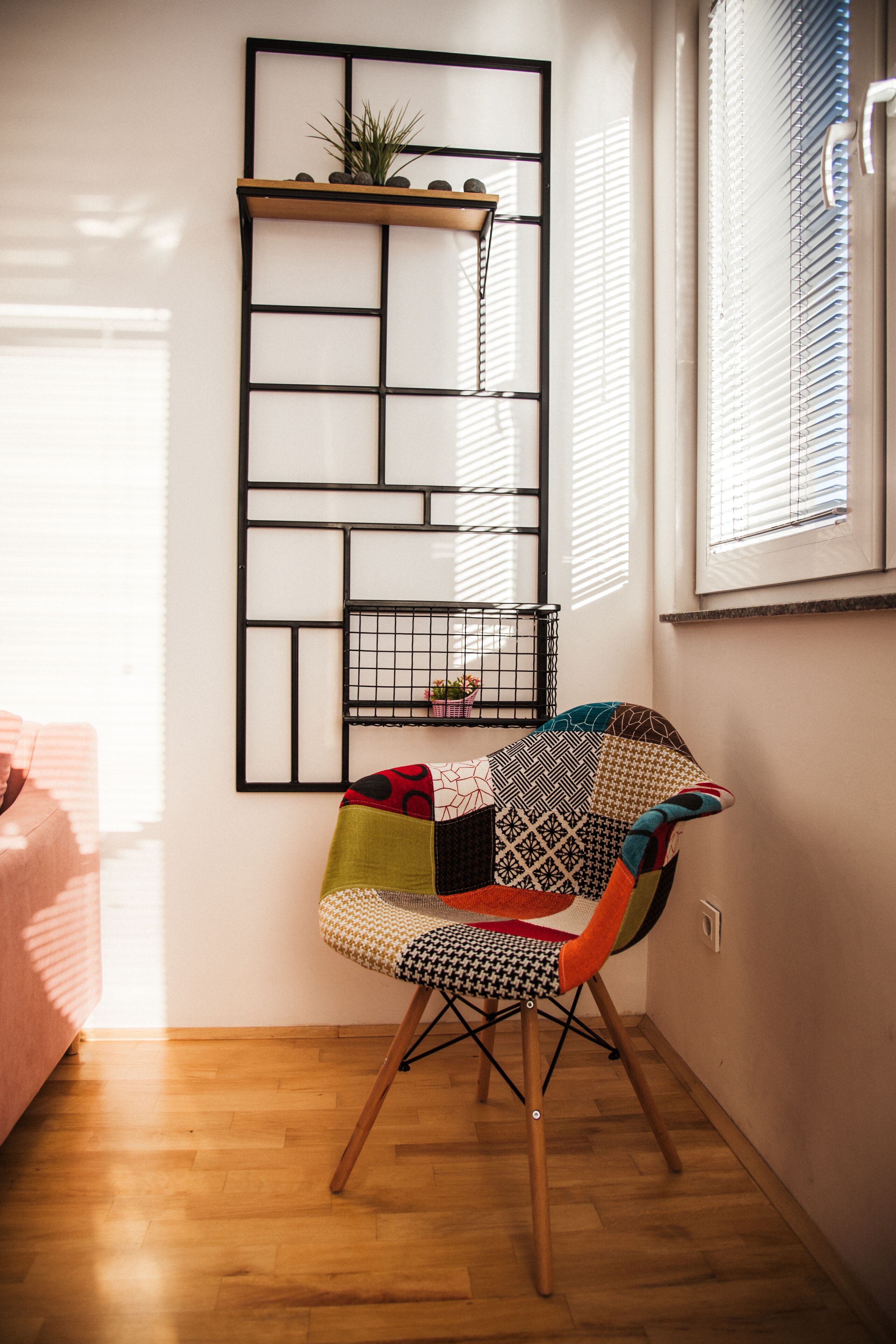 Une pièce avec un plancher en bois, une chaise à motifs, une étagère en métal noir ornée de plantes et une fenêtre avec des stores.