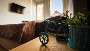 Een kamer met een bed, een klein tafeltje met een speelgoedfiets en een bloempot, een televisie aan de muur, en een raam met uitzicht op een huis.
