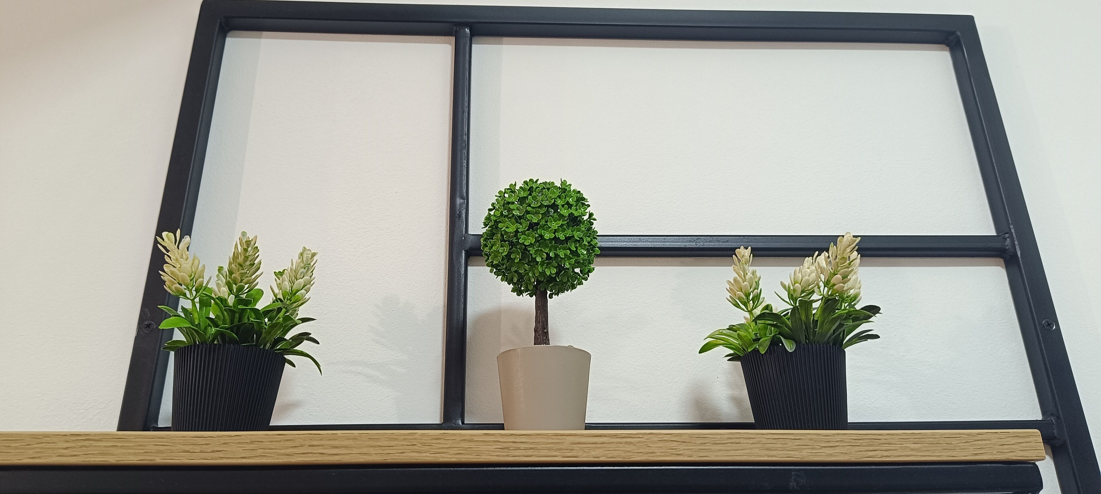 A window with a black frame and a shelf below it. On the shelf, there are three potted plants, one in a black pot and two in beige pots.