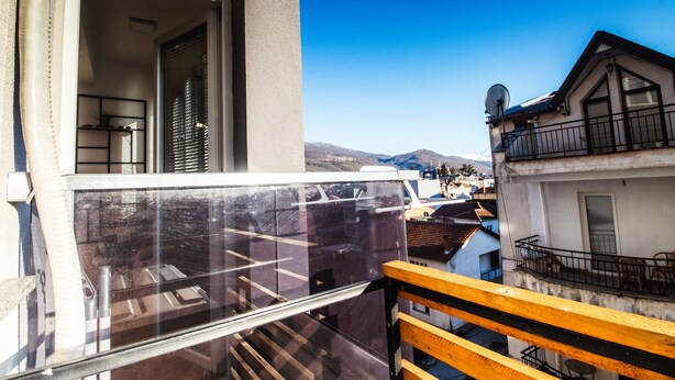 Een balkon met houten hekwerk, een glazen deur en uitzicht op een stad met gebouwen en bergen op de achtergrond.