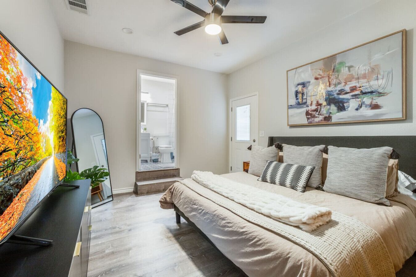 A bedroom with a bed, a ceiling fan, a painting, and a TV on a stand.