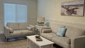 A living room with two beige sofas, a white coffee table, a lamp, and a painting of a beach scene.
