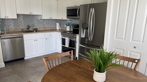 A kitchen with white cabinets, a stainless steel refrigerator, a stove, and a sink. There is a wooden dining table with chairs and a potted plant.