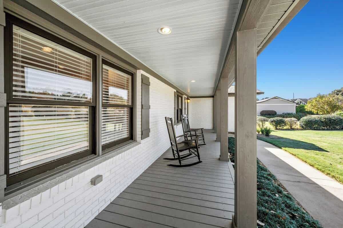 A covered porch with a brick wall, large windows with blinds, and a wooden deck.