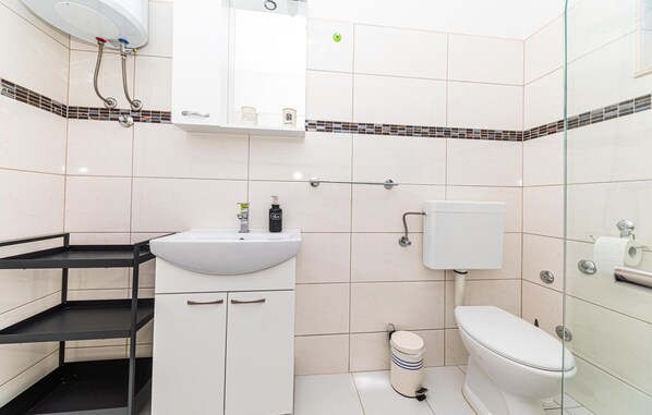 Une salle de bain avec un lavabo, des toilettes et une douche, le tout de couleur blanche. On y trouve des étagères noires et une paroi de douche en verre.