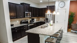 A modern kitchen with dark cabinets, granite countertops, stainless steel appliances, and a bar area with stools. - Luxury 5bd townhome | Gated Resort Community (Kissimmee)