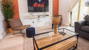 A living room with a flat-screen TV mounted on the wall displaying "NETFLIX", a brown chair, a glass coffee table, a blue ottoman, and a grey sofa. - Luxury 5bd townhome | Gated Resort Community (Kissimmee)