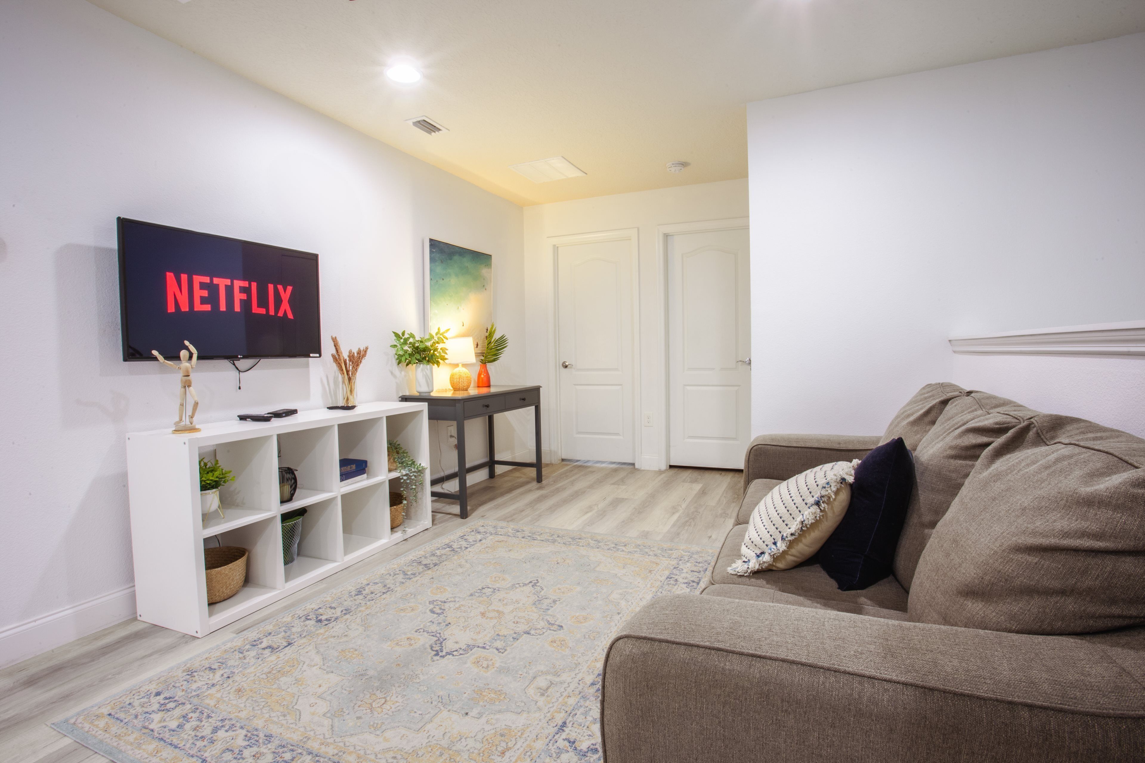 Ein Wohnzimmer mit einem Flachbildfernseher, der 'NETFLIX' anzeigt, einem weißen Konsolentisch mit Dekorationen, einem braunen Sofa und einem gemusterten Teppich.
