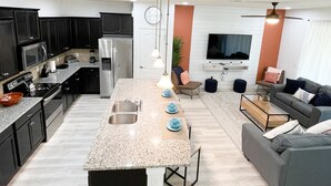 A modern kitchen with dark cabinets, a central island, and a dining area with a sofa and coffee table. - Luxury 5bd townhome | Gated Resort Community (Kissimmee)