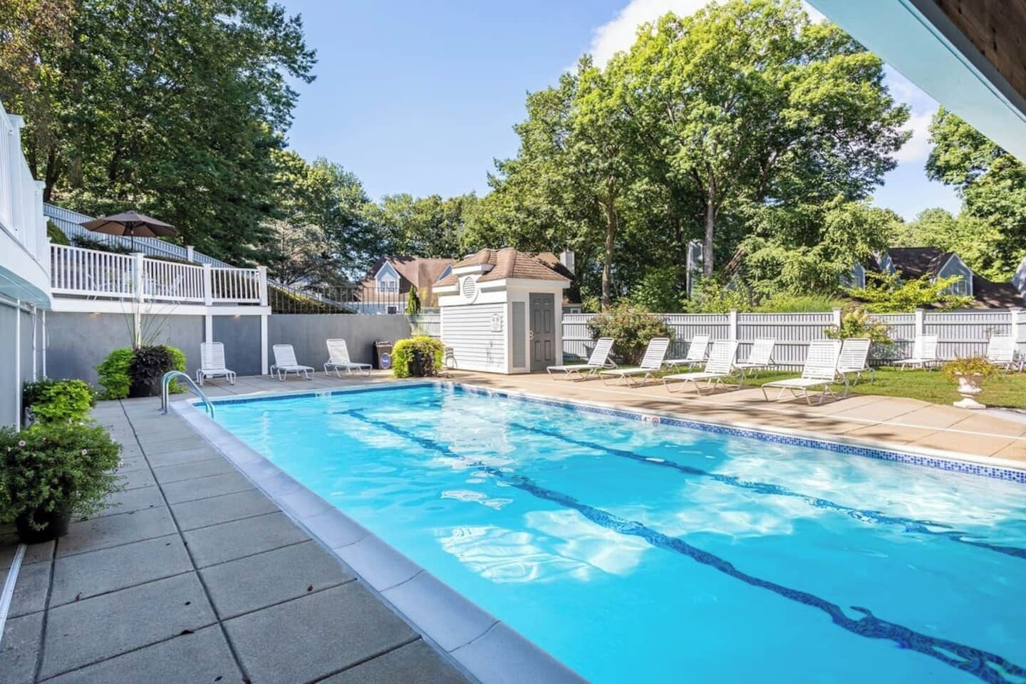 A swimming pool with clear blue water, surrounded by a white fence, and a covered patio area with lounge chairs.