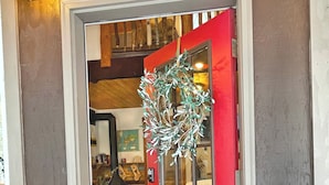 Eine rote Tür mit einem 'WELCOME'-Schild darüber, ein Kranz hängt an der Tür, und ein Holzinnenraum ist hindurch sichtbar.