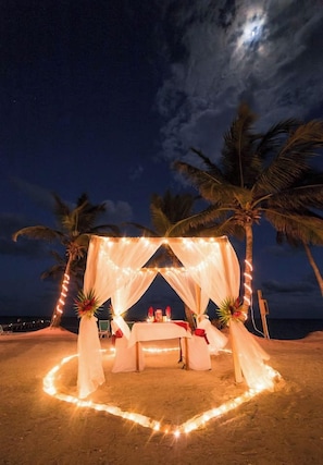 A beach setting with a canopy and string lights, palm trees, and a table with chairs.