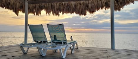 Une terrasse en bois avec deux chaises longues, située sous un toit de chaume et offrant une vue sur une mer calme.