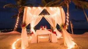 Un décor de plage avec un auvent et des guirlandes lumineuses, des palmiers et une table décorée de bougies.