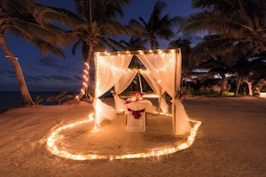 A beach setting with a canopy structure adorned with lights, a table with a red ribbon, and palm trees in the background.