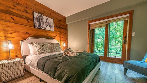A wooden-paneled bedroom with a bed, two bedside lamps, a large window, and a view of greenery outside. - Chimney Hill - 02) First-Floor Suite 2 (Lambertville)