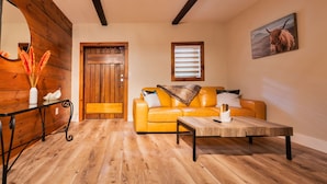 A cozy living room with a wooden floor, a yellow sofa, a coffee table, and a wooden door. - Chimney Hill - 02) First-Floor Suite 2 (Lambertville)