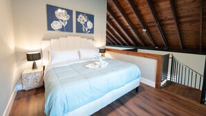 Un dormitorio con techo de madera, una cama con ropa de cama blanca, dos lámparas fijadas a la pared y dos cuadros con motivos florales.